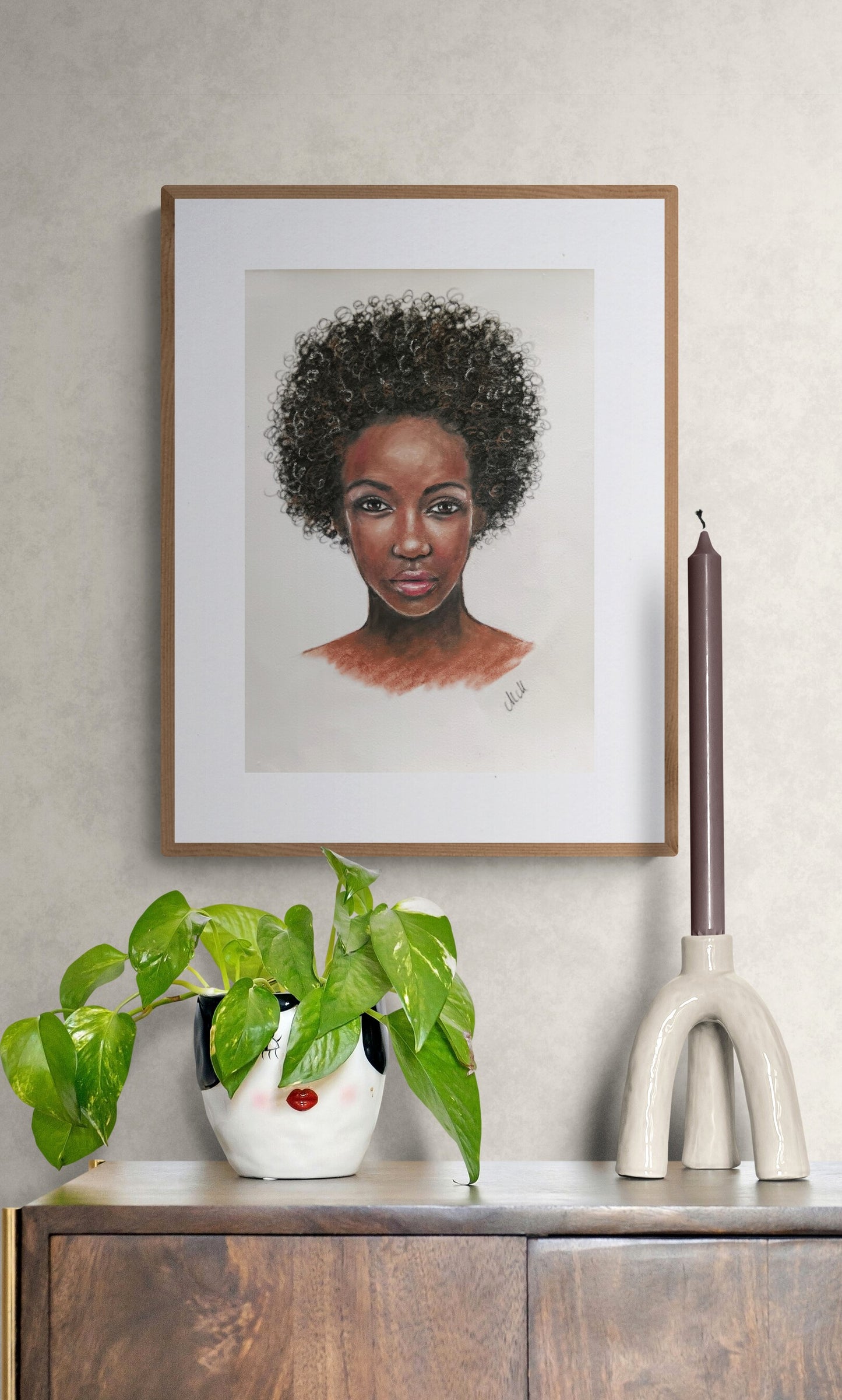 Framed portrait of a woman with an afro above a plant and decorative items on a shelf.