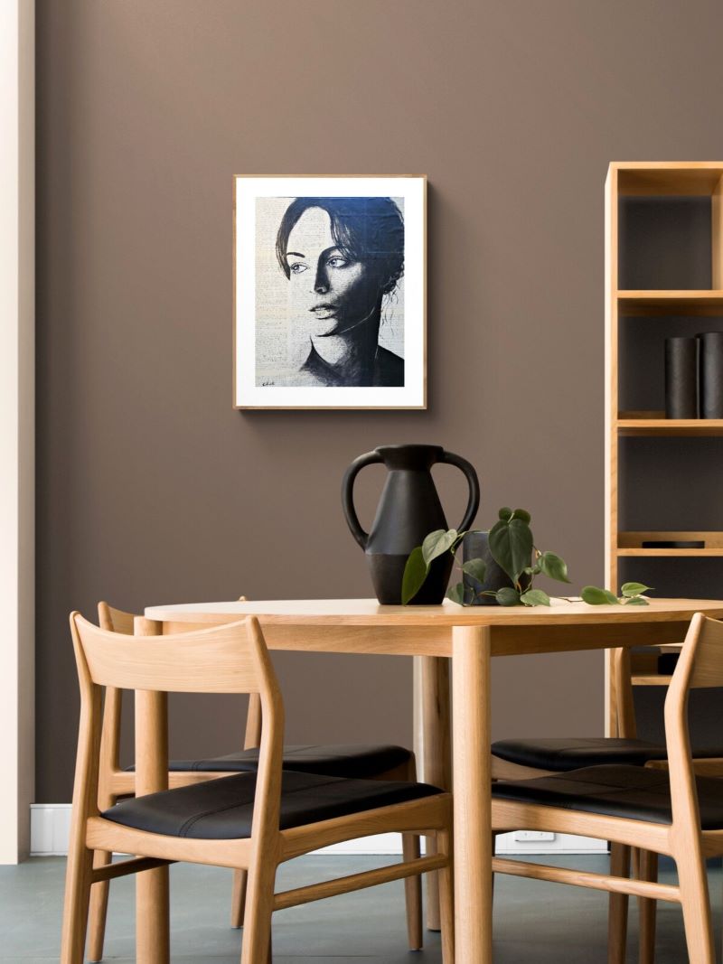 A framed black and white portrait print of a woman is displayed on a brown wall in a modern dining room setting with a wooden table, chairs, and a black pitcher.
