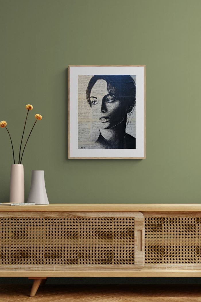 A framed black and white portrait print of a woman with dark hair, displayed on a sage green wall above a light wood cabinet with a woven cane front. Two white vases with yellow flowers sit on the cabinet.