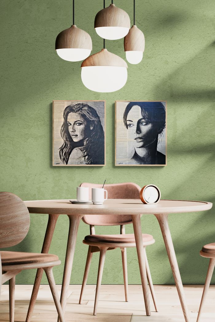 A styled dining room corner with a round light wood table, pink upholstered chairs, three hanging pendant lights, and two framed black-and-white portrait prints on a textured sage green wall.