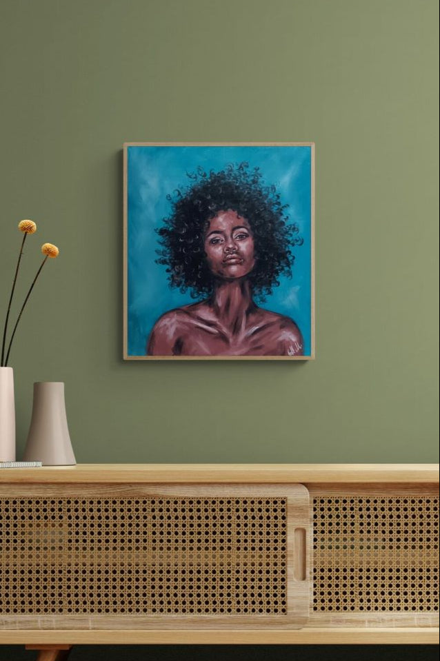 A framed portrait painting of a Black woman with voluminous curly hair against a bright blue background, displayed on a sage green wall above a light wood cabinet with a woven front.