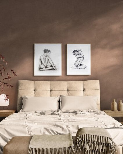 A styled bedroom scene with a bed dressed in neutral beige and cream bedding, a tufted beige headboard, two framed black-and-white nude figure artworks on the wall, and a warm brown wall behind.