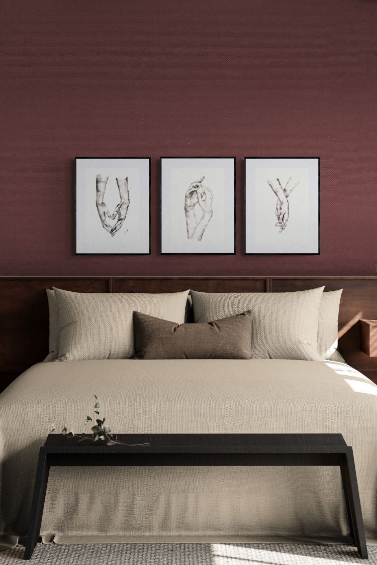 A styled bedroom scene with a bed dressed in beige bedding and pillows, beneath three framed minimalist line-art prints on a deep burgundy wall. A dark wood bench sits at the foot of the bed.