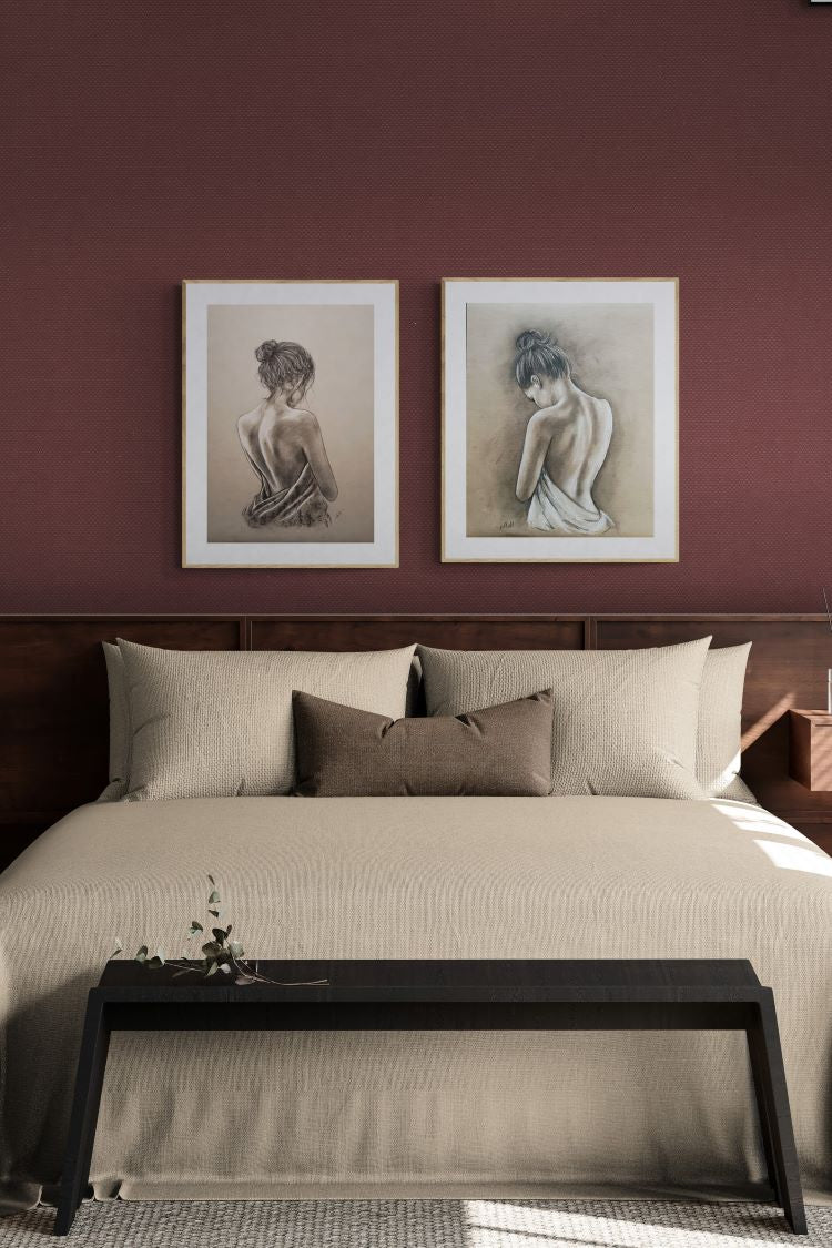 A styled bedroom scene with a neatly made bed in cream bedding, a dark wood headboard and bench, two framed monochrome sketches on a deep burgundy wall, and a textured rug.