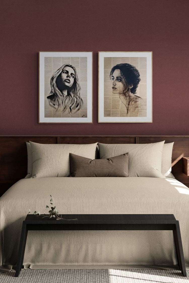 A styled bedroom scene with a bed dressed in beige bedding, two framed monochrome portrait prints hanging on a dark burgundy wall above the headboard, and a black bench at the foot of the bed.