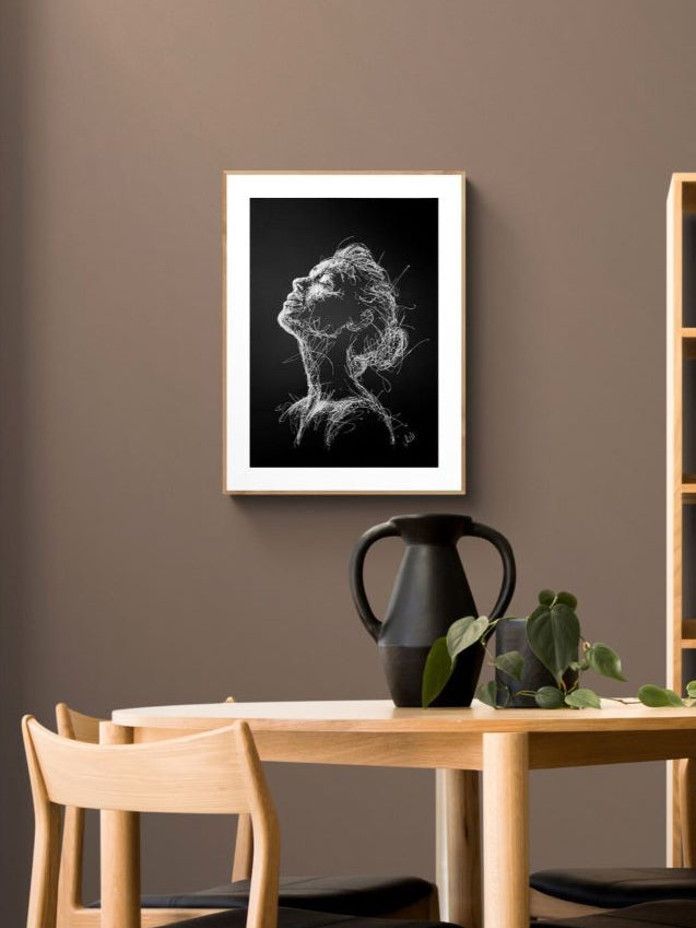 A framed black and white sketch portrait of a woman's head and neck hanging on a brown wall above a light wood dining table with chairs.