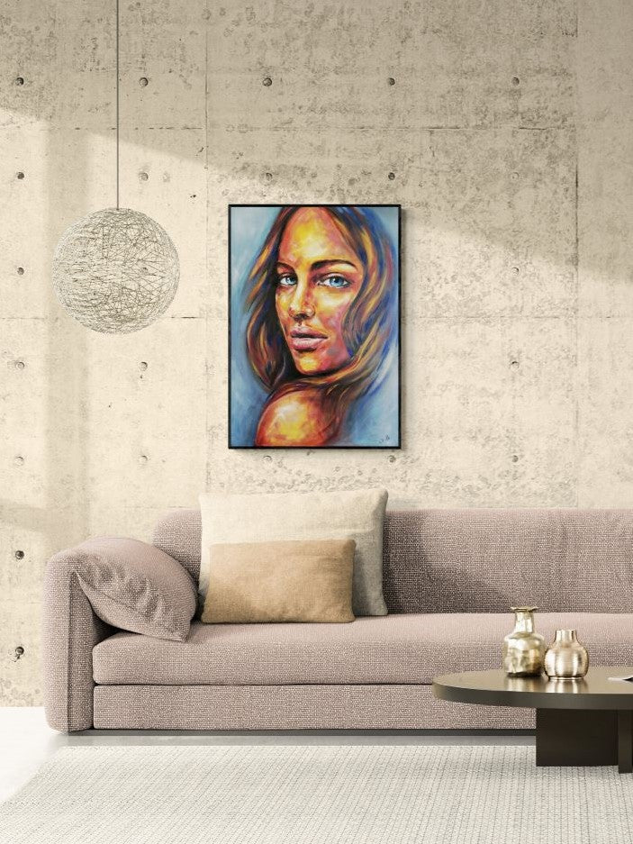 A framed portrait painting of a woman with warm skin tones and blue hair, displayed on a concrete-textured wall above a beige sofa in a modern living room setting.