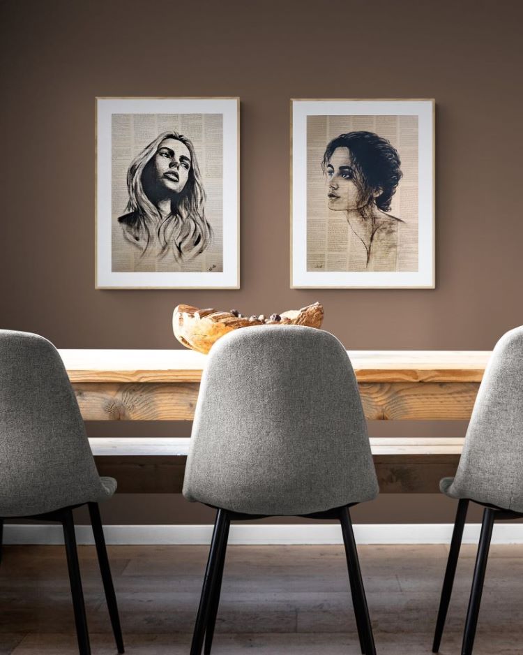Two framed black and white portrait sketches of women displayed side-by-side on a brown wall above a wooden dining table with three gray chairs.