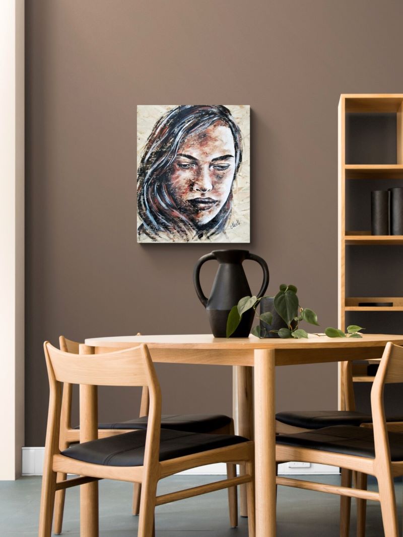 A square canvas art print featuring a stylized portrait of a woman, displayed on a brown wall in a modern dining room setting above a wooden table and chairs.