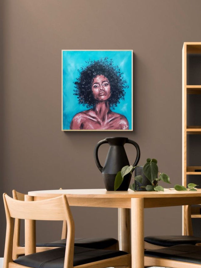 A framed portrait painting of a Black woman with voluminous dark hair against a bright turquoise background, displayed on a brown wall above a light wood dining table and chairs.