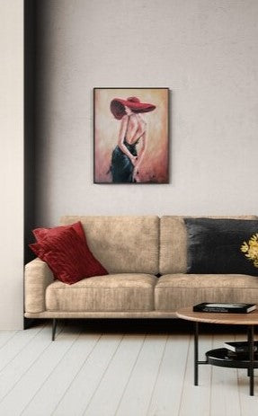 A framed vertical art print depicting a woman in a red hat and dark dress, displayed on a light gray wall above a beige sofa with red and black pillows in a modern living room setting.