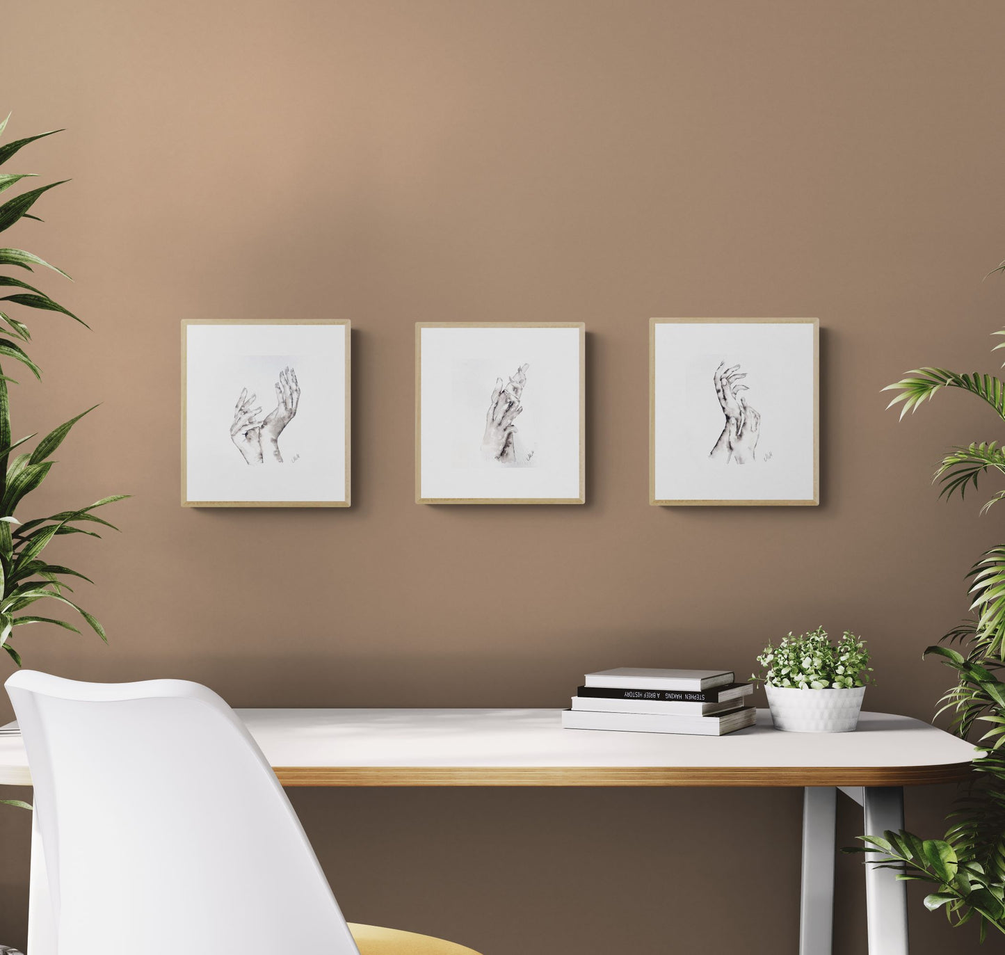 Three framed artworks on a brown wall above a desk with books and a plant.