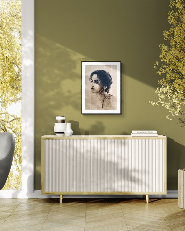 A framed art print featuring a sepia-toned portrait of a woman, displayed on an olive green wall above a ribbed white and gold sideboard in a modern, sunlit room.