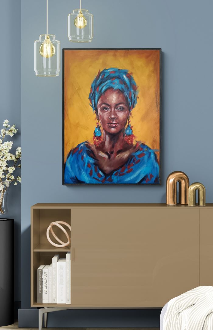 A framed portrait painting of a woman with a blue headwrap and blue garment against a warm yellow-orange background, displayed on a blue wall above a beige cabinet in a styled interior.