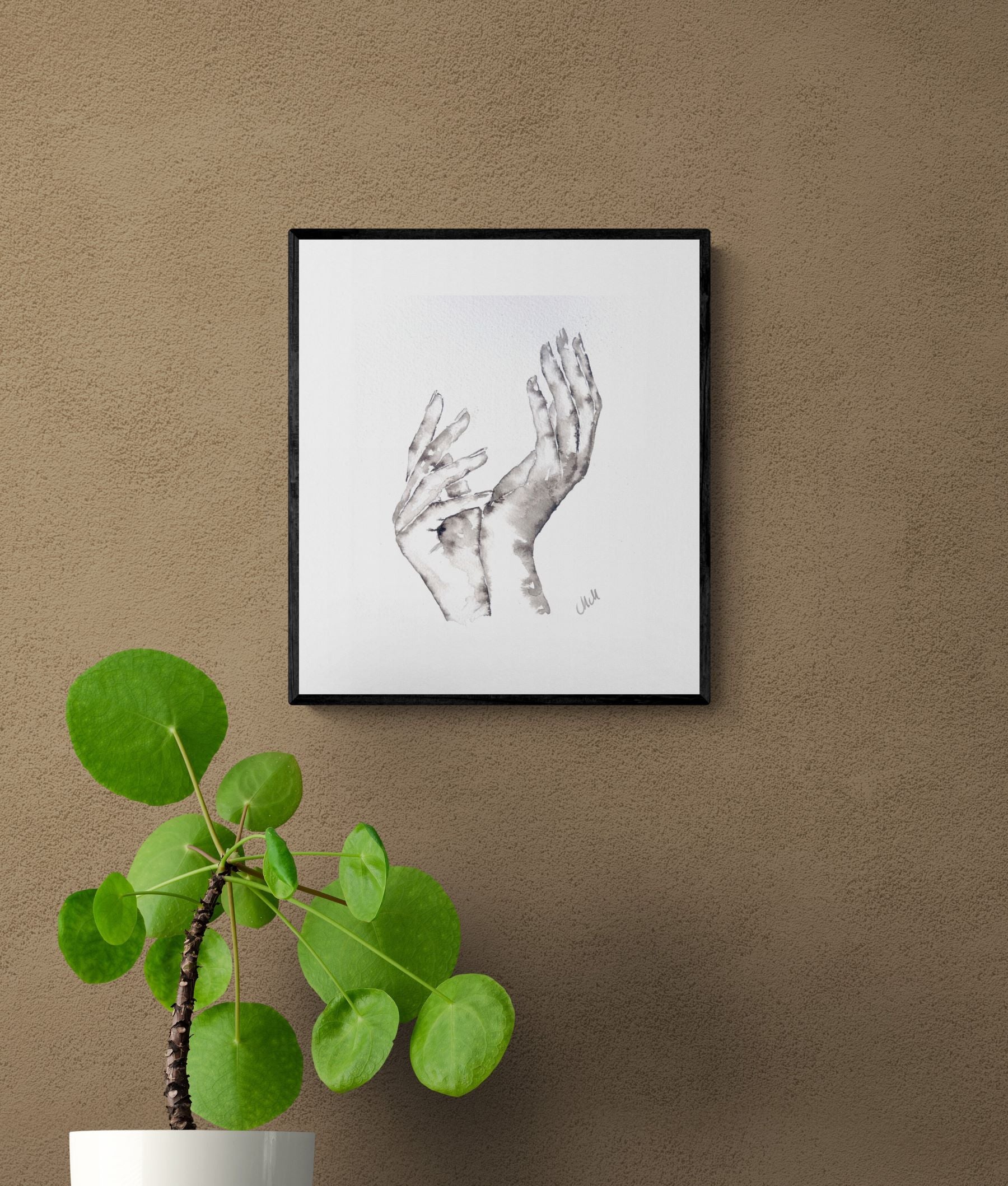 Framed artwork of two hands on a brown wall with a plant below.