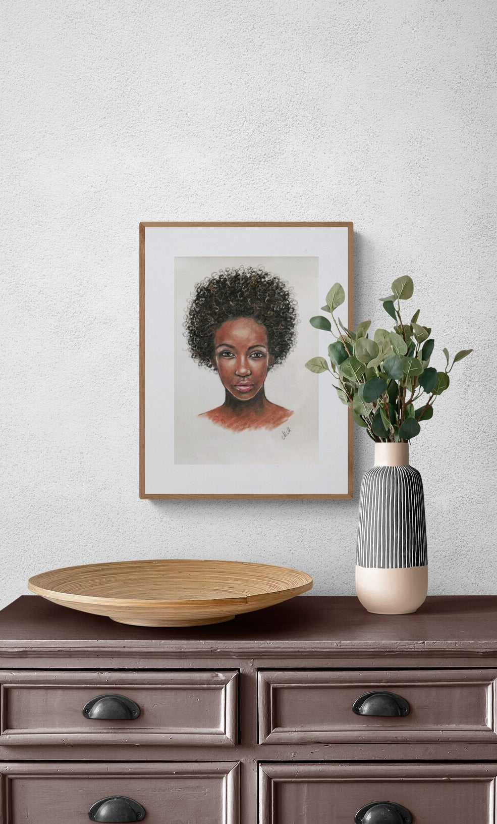 Framed portrait of a woman on a wall above a wooden dresser with a vase and bowl.